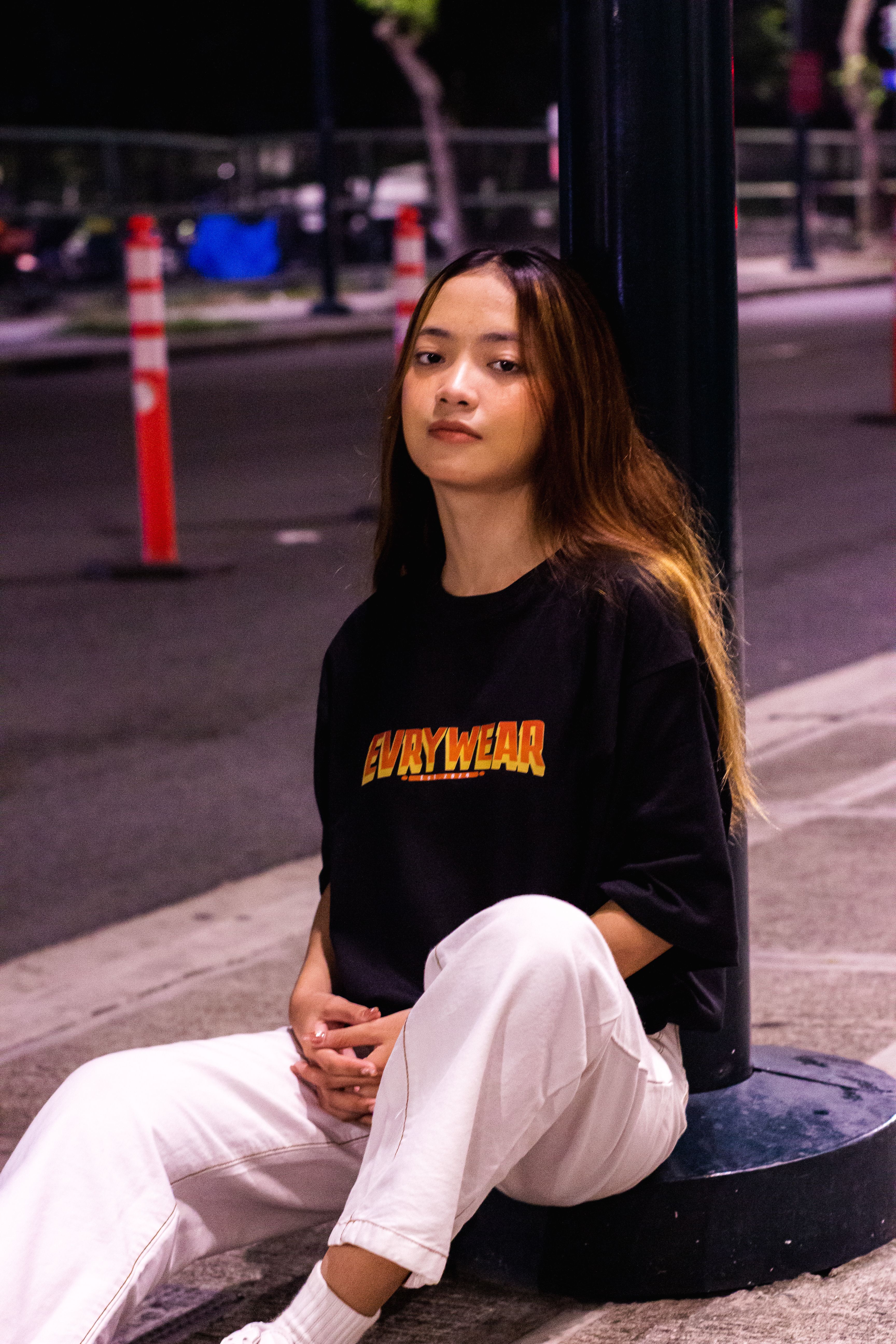 Stylish young woman sitting by a streetlight at night in a city environment, exuding casual urban fashion.