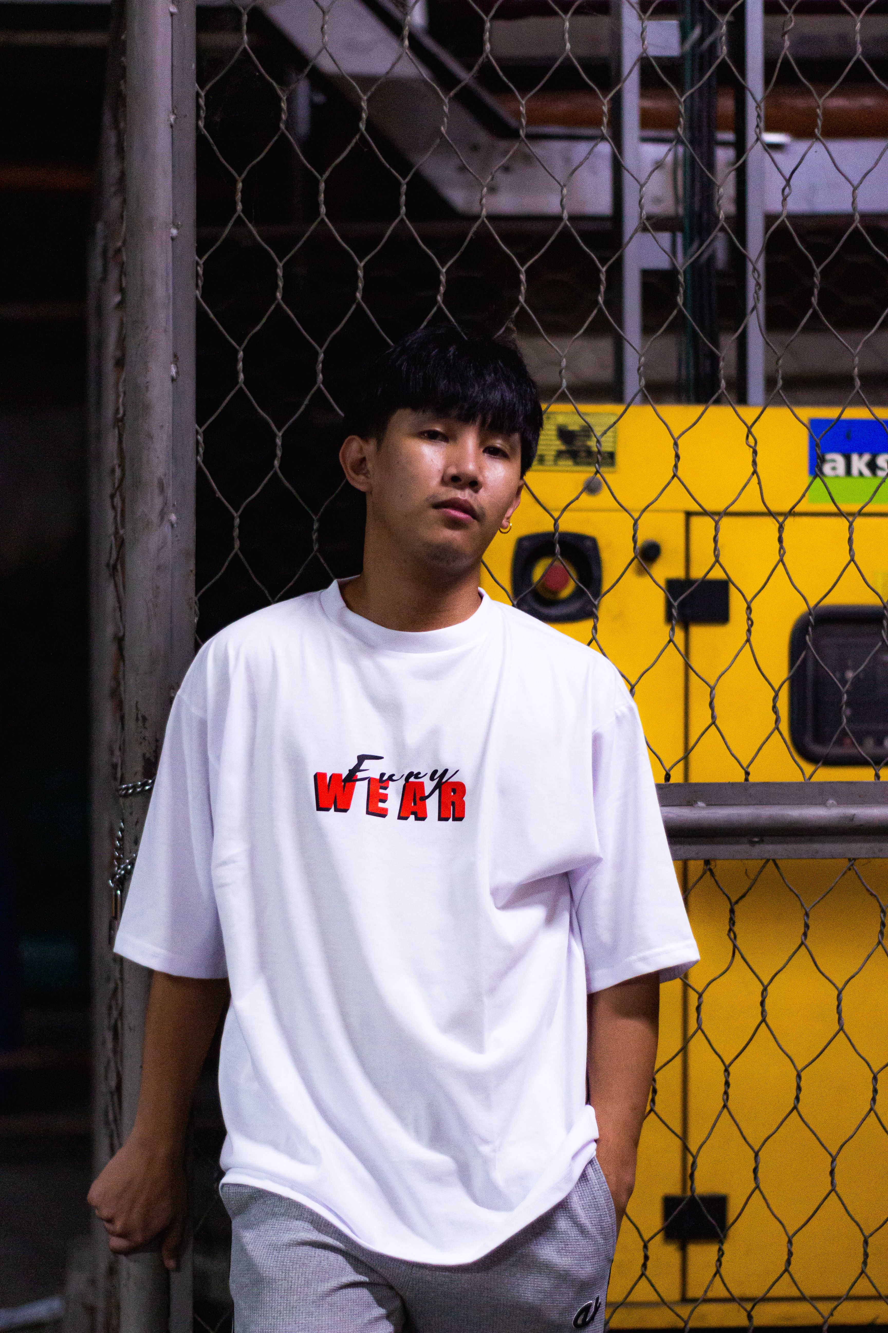 Young man in a white t-shirt posing casually indoors with industrial backdrop.