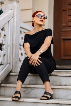 Fashionable woman with sunglasses sitting outdoors in a black outfit.