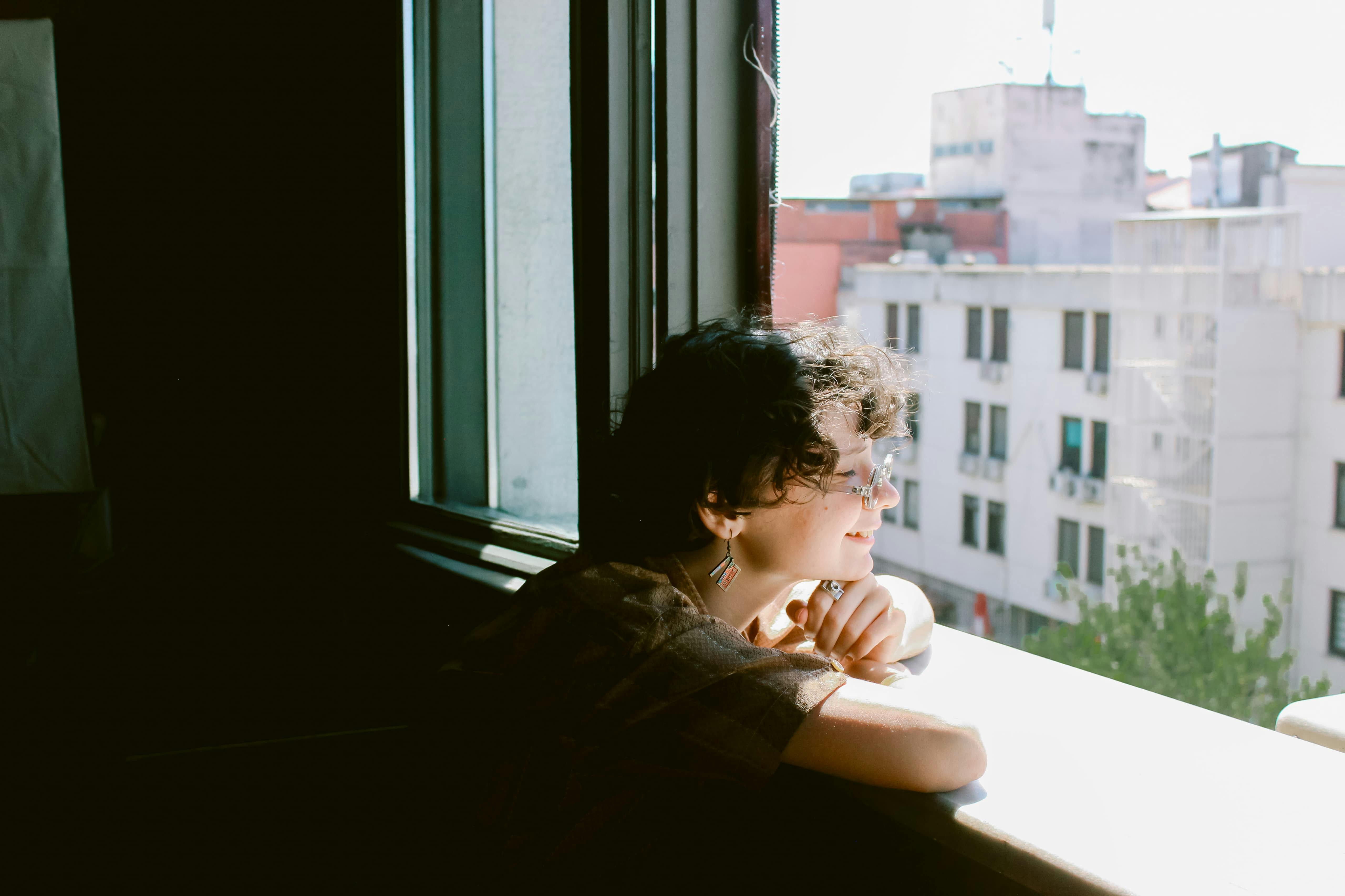 Free A young woman leans on a windowsill, enjoying sunlight with a cityscape view in the background. Stock Photo