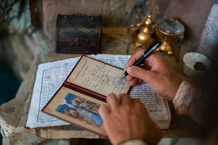 Person Writing In Small Diary Book With Religious Picture