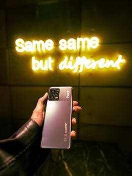 Hand holding a Poco smartphone with a glowing neon sign reading 'Same same but different.'