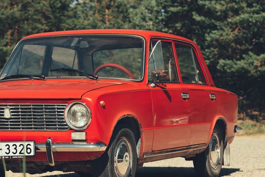 A vibrant red vintage Lada car parked outdoors in Saulkrasti, Latvia, against a green forest background.