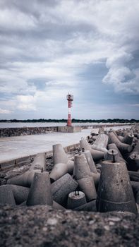 Concrete tetrapods and a red lighthouse against a cloudy sky by the coast.