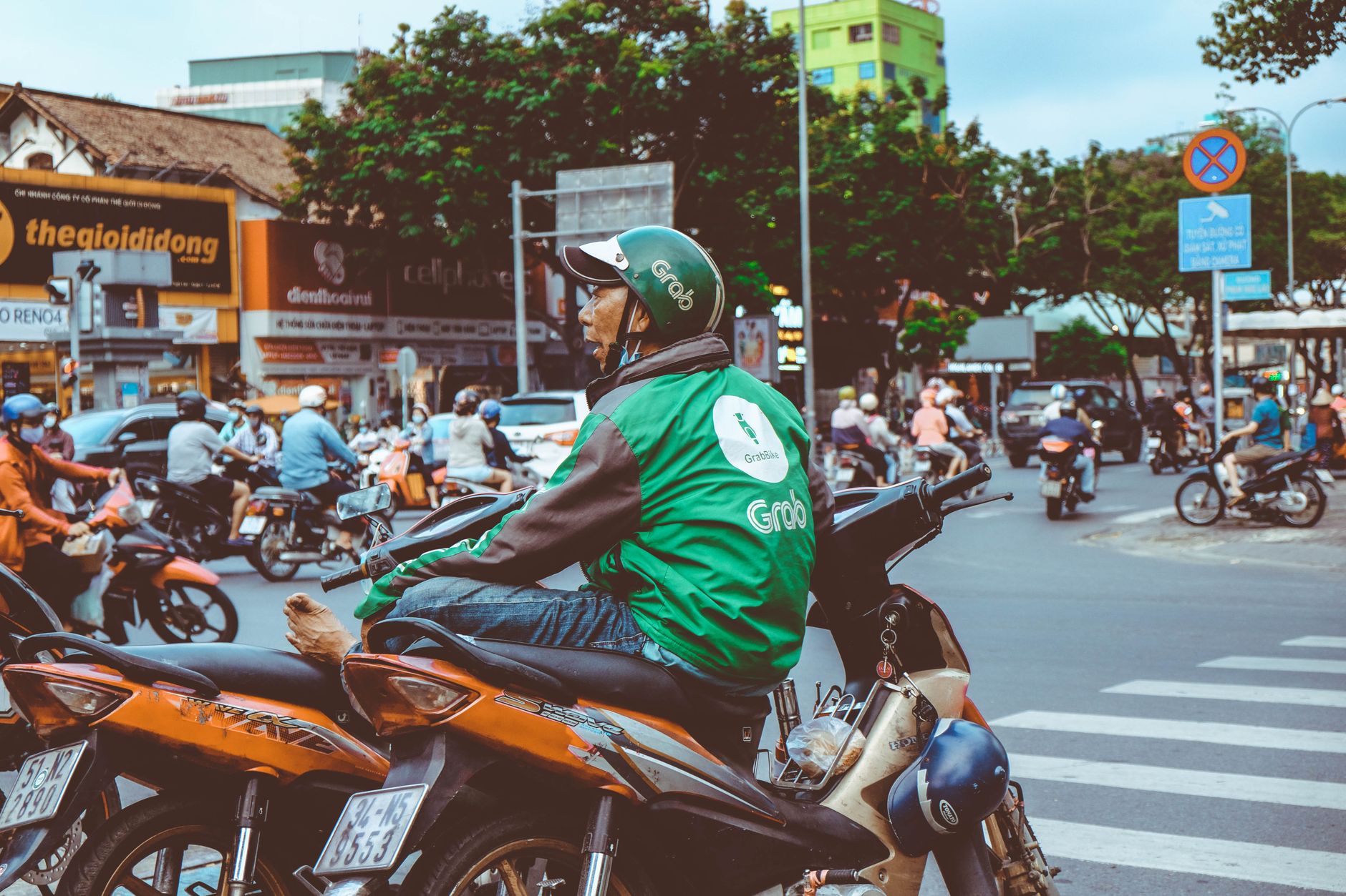 Motorbike rider in green jacket waits at a bustling urban intersection, surrounded by traffic.