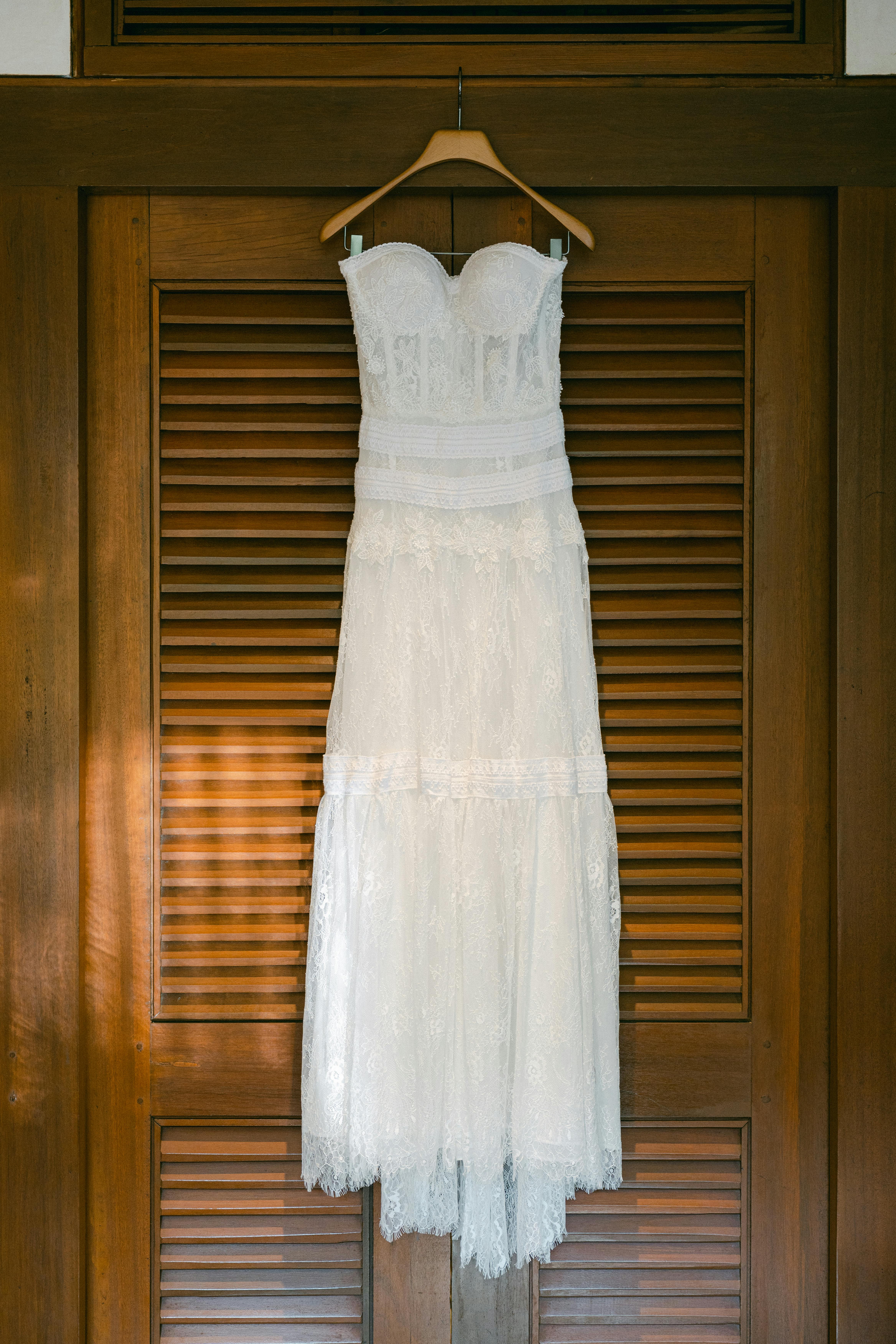 A beautiful white wedding dress hangs gracefully on a wooden hanger indoors.
