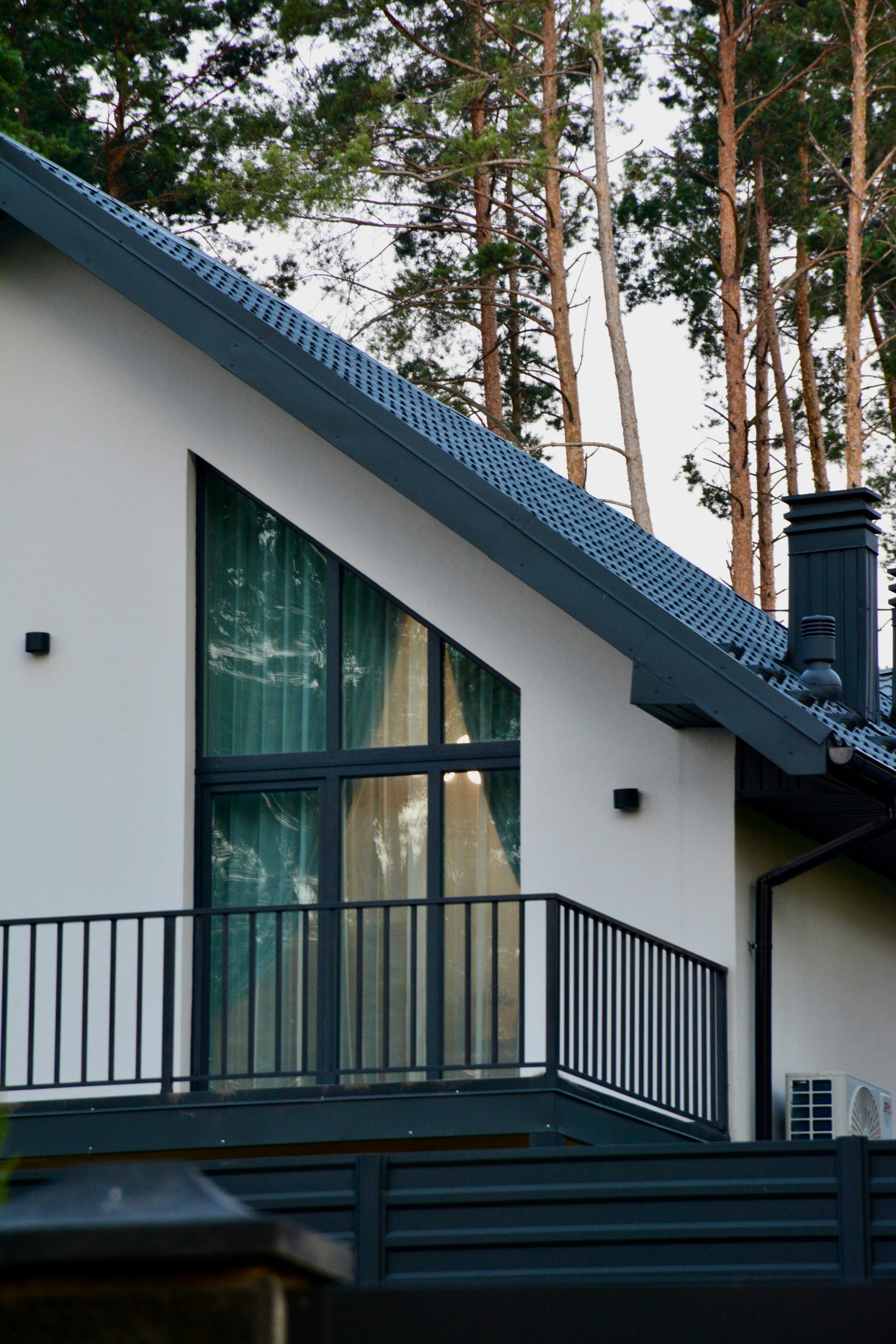 A modern house featuring a large window and balcony amidst tall pine trees.
