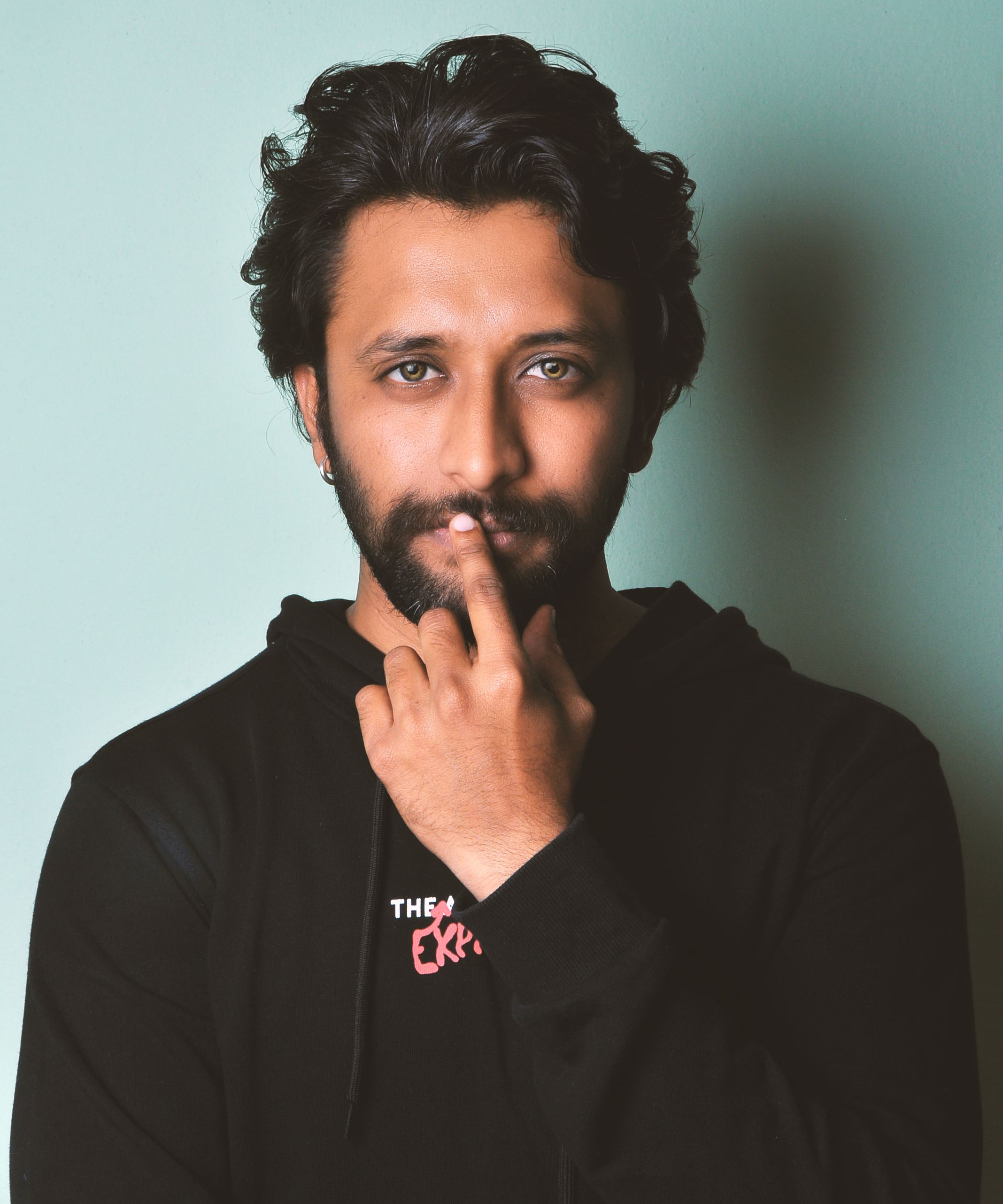 Portrait of a thoughtful man in a black hoodie, against a green background, with a finger on his lips.