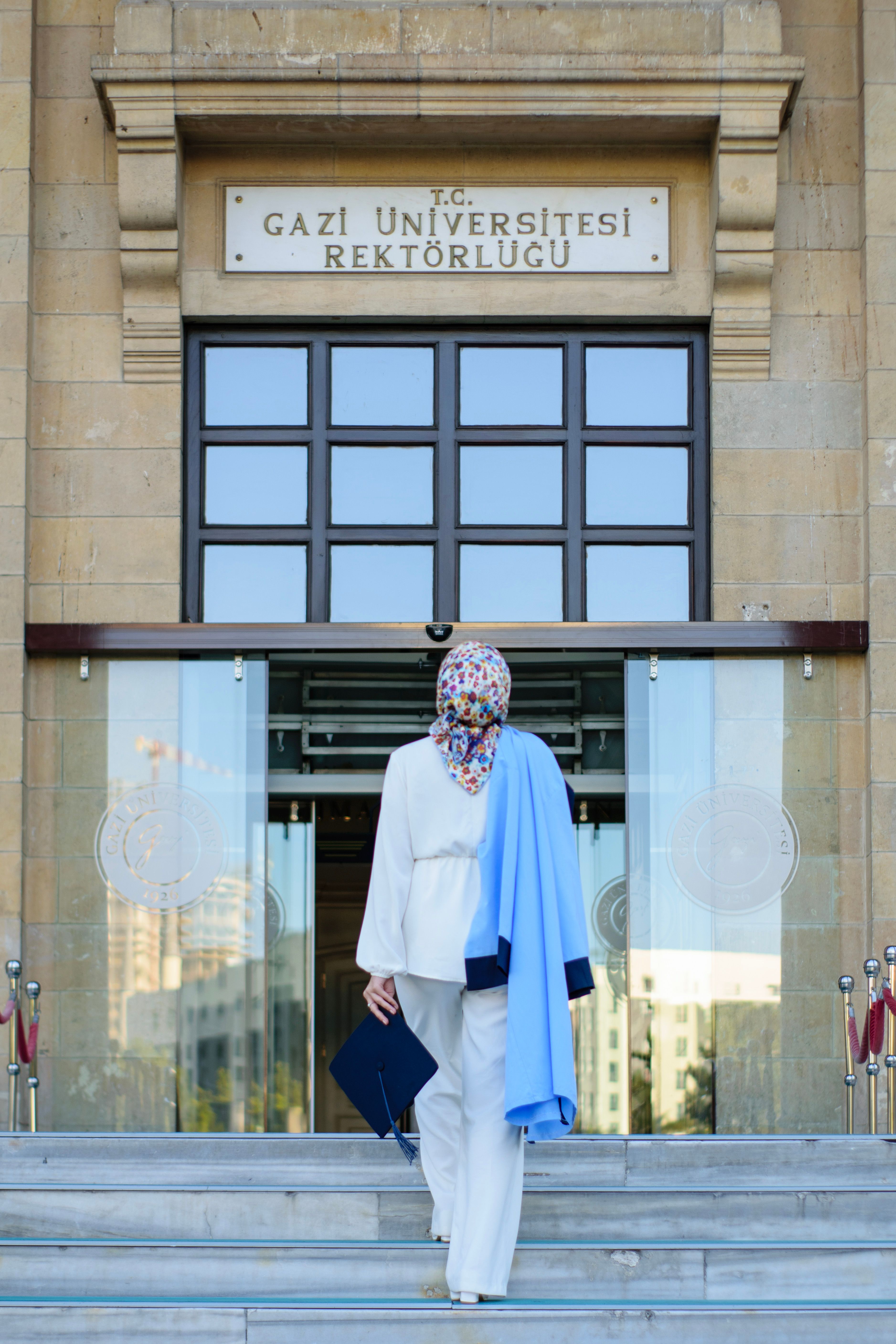 Woman in headscarf holding diploma at Gazi University entrance.