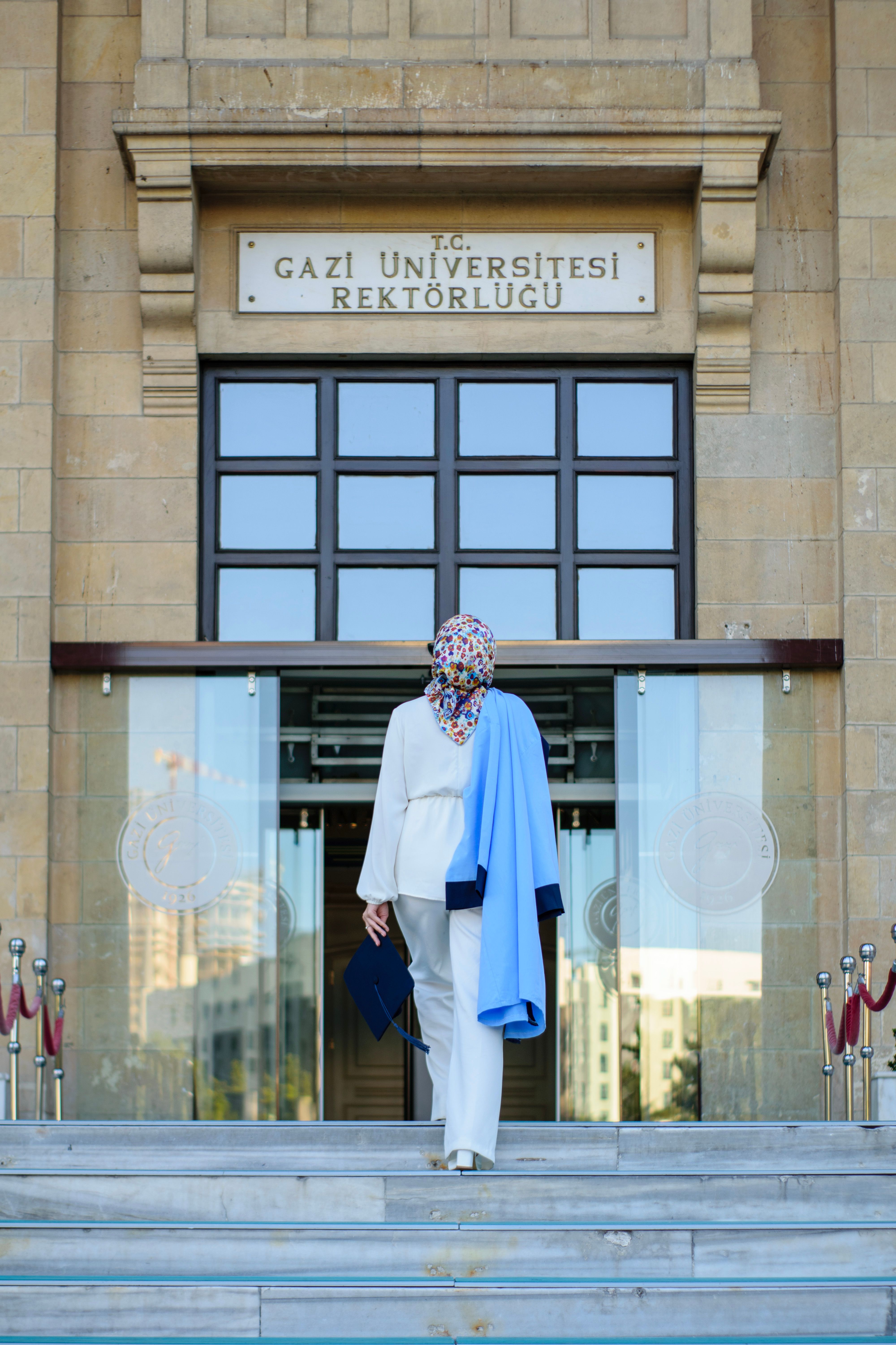 A woman in a hijab walking towards Gazi University's entrance on graduation day.