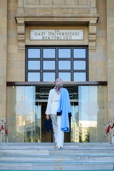 A woman in a hijab walking towards Gazi University's entrance on graduation day.