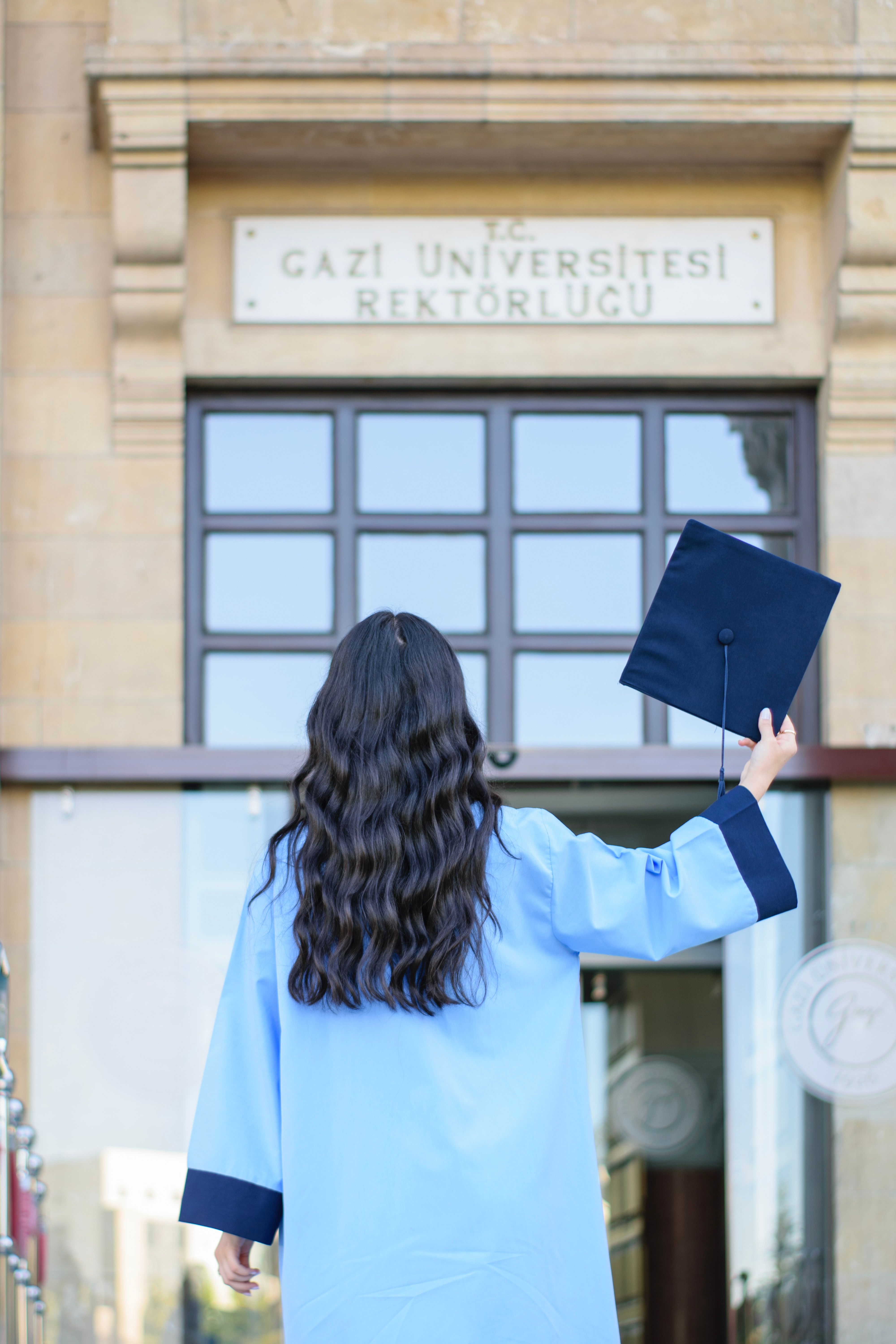Back view of a woman holding a diploma, celebrating graduation at Gazi University entrance.