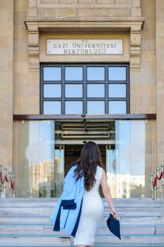 Woman in graduation attire walking towards a university entrance with diploma in hand.