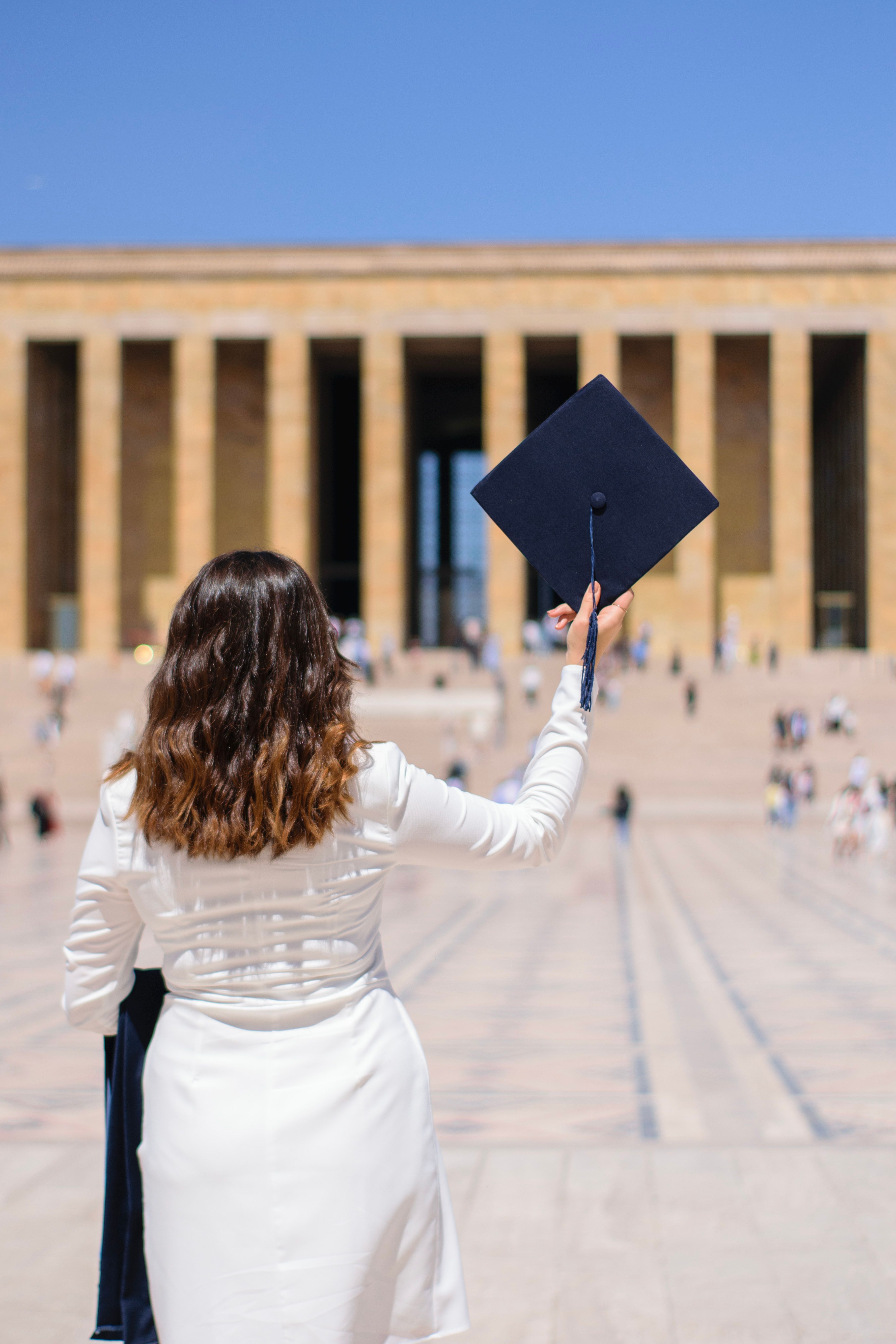 Female graduate in white celebrating success at a historic monument, holding a mortarboard.