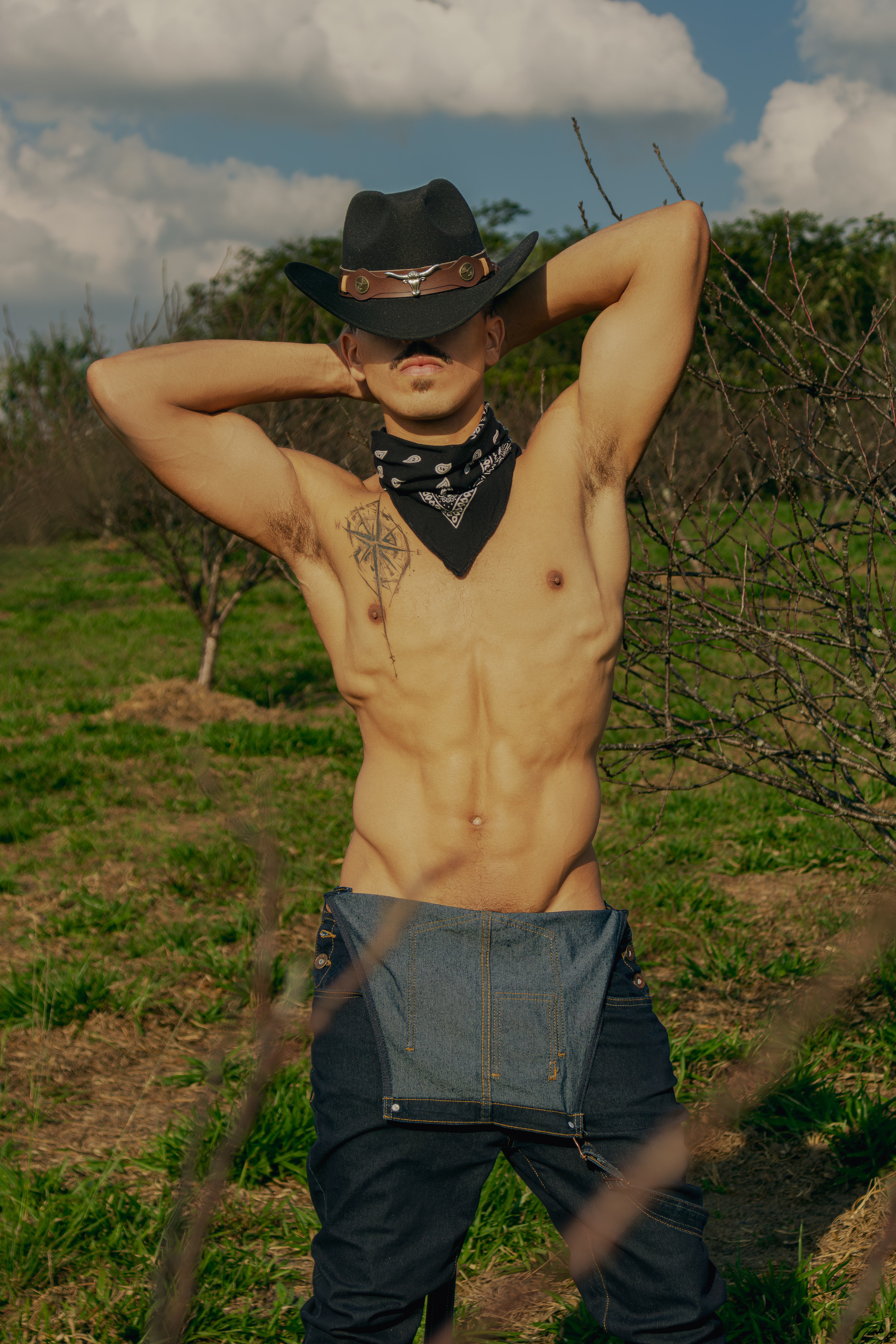 A shirtless cowboy wearing a bandana and hat poses confidently in a rural field.