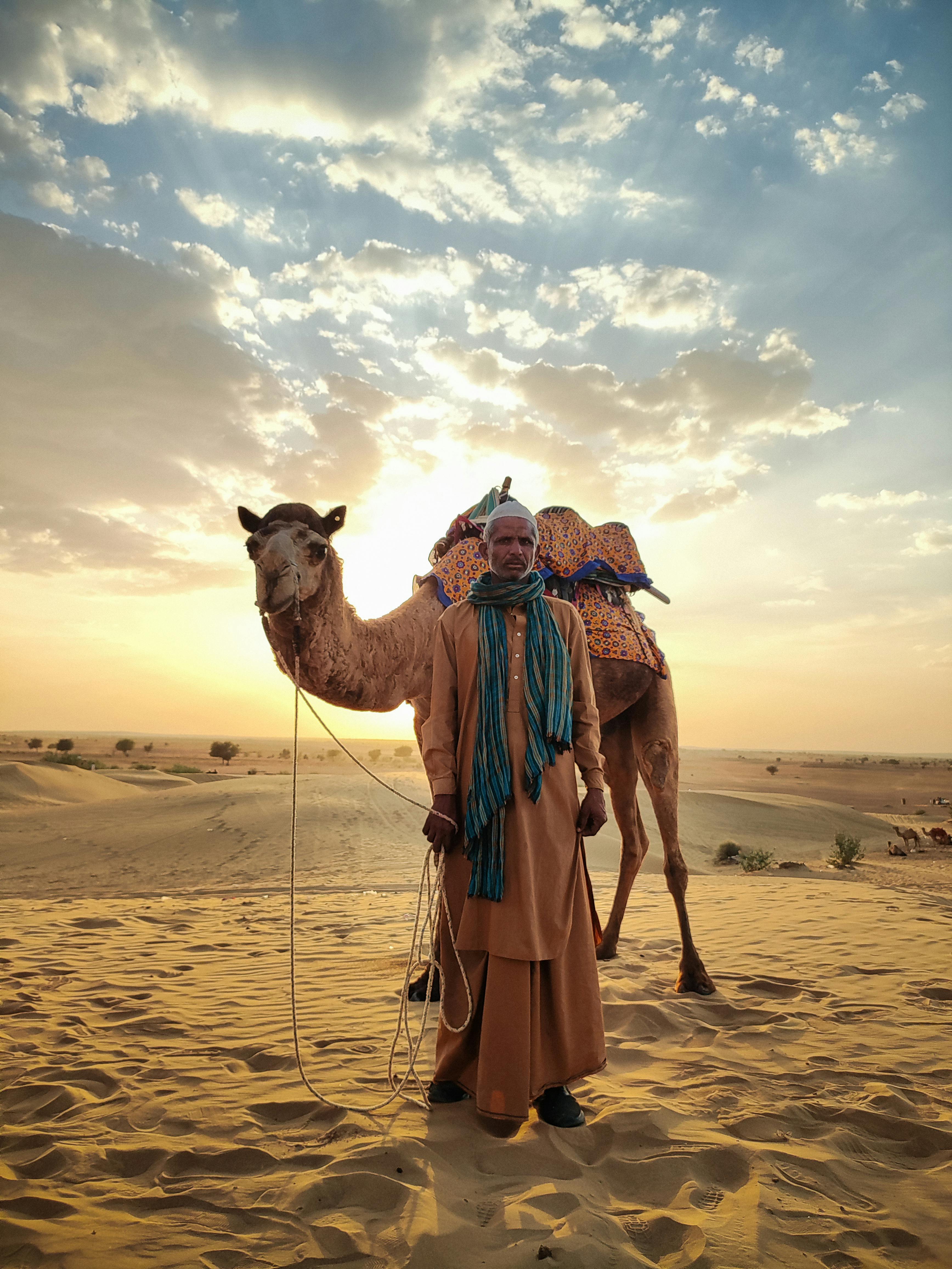 man in traditional clothing posing with a camel in a desert