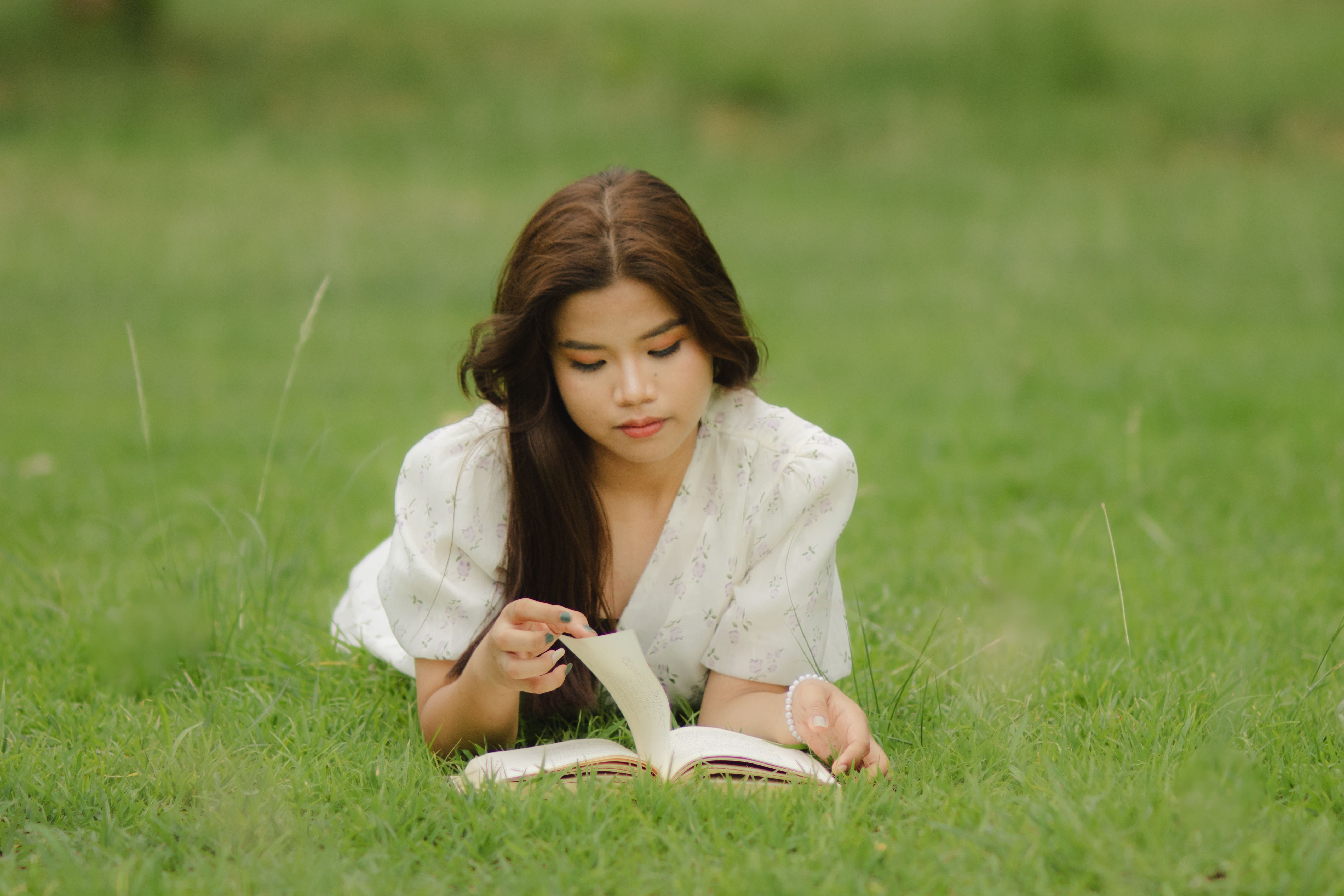 A serene portrait of a young woman lying in a green field, absorbed in reading a book.