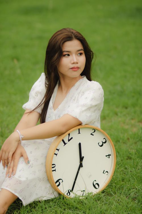 Free Portrait of a young woman in a white dress sitting on grass with a large clock. Stock Photo