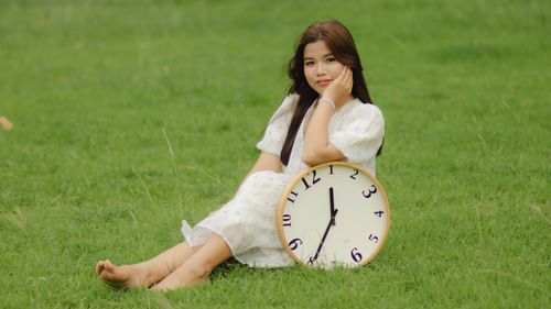 Free Asian woman in white dress sitting barefoot on grass holding clock, outdoor setting. Stock Photo