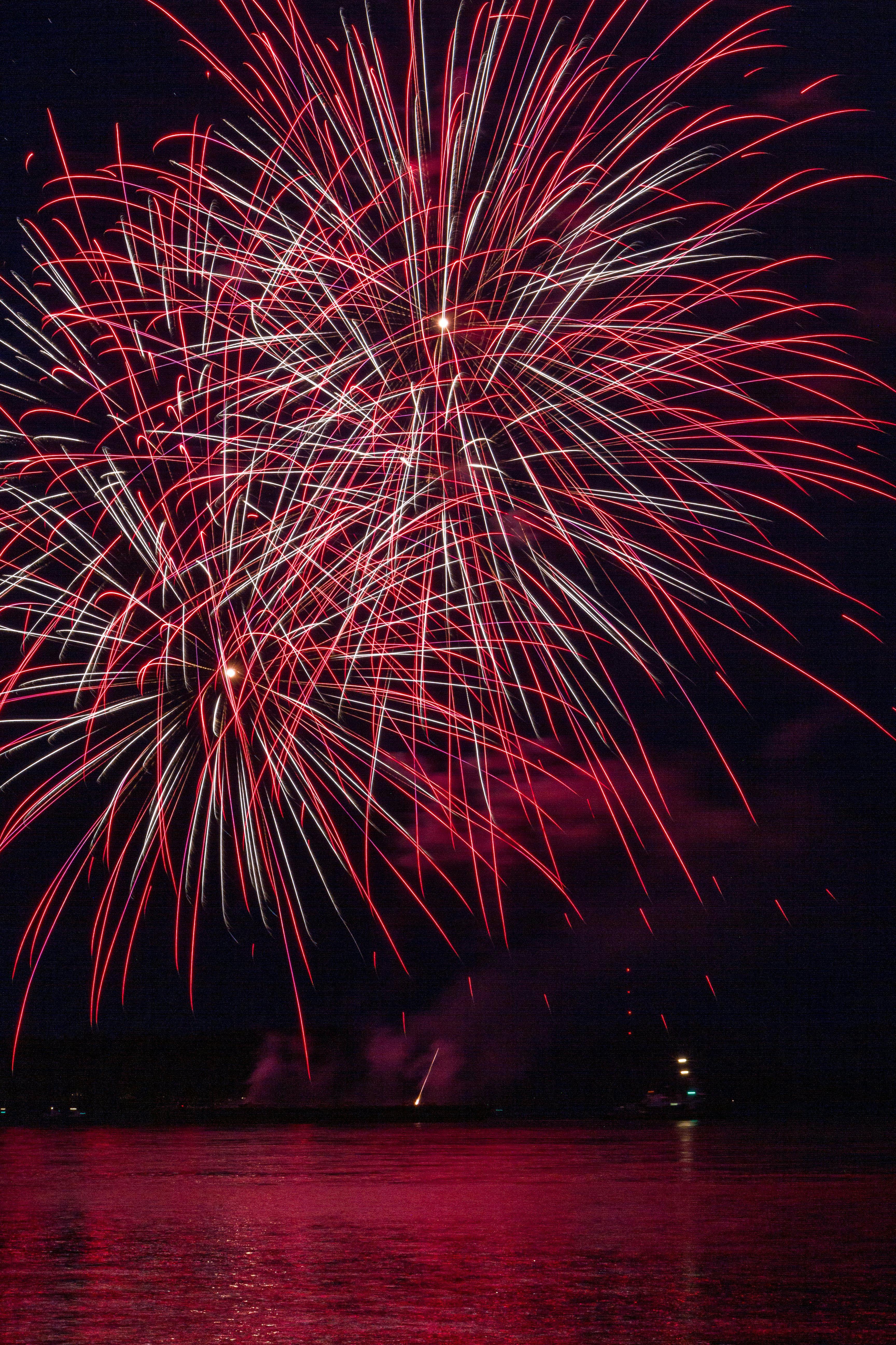 Stunning red fireworks burst over a serene body of water during a night festival celebration.