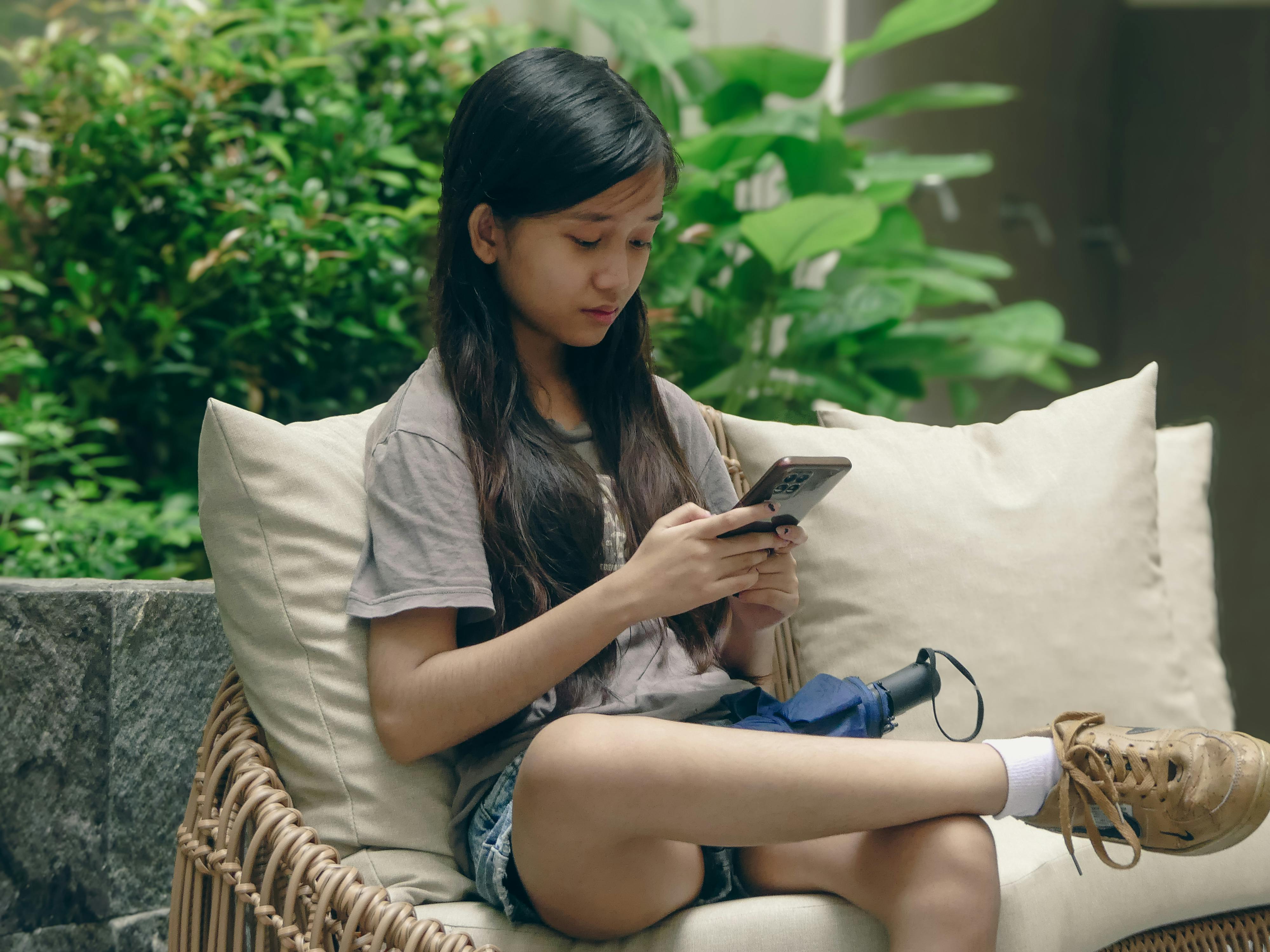 Asian girl sitting outdoors, using a smartphone, surrounded by lush greenery.