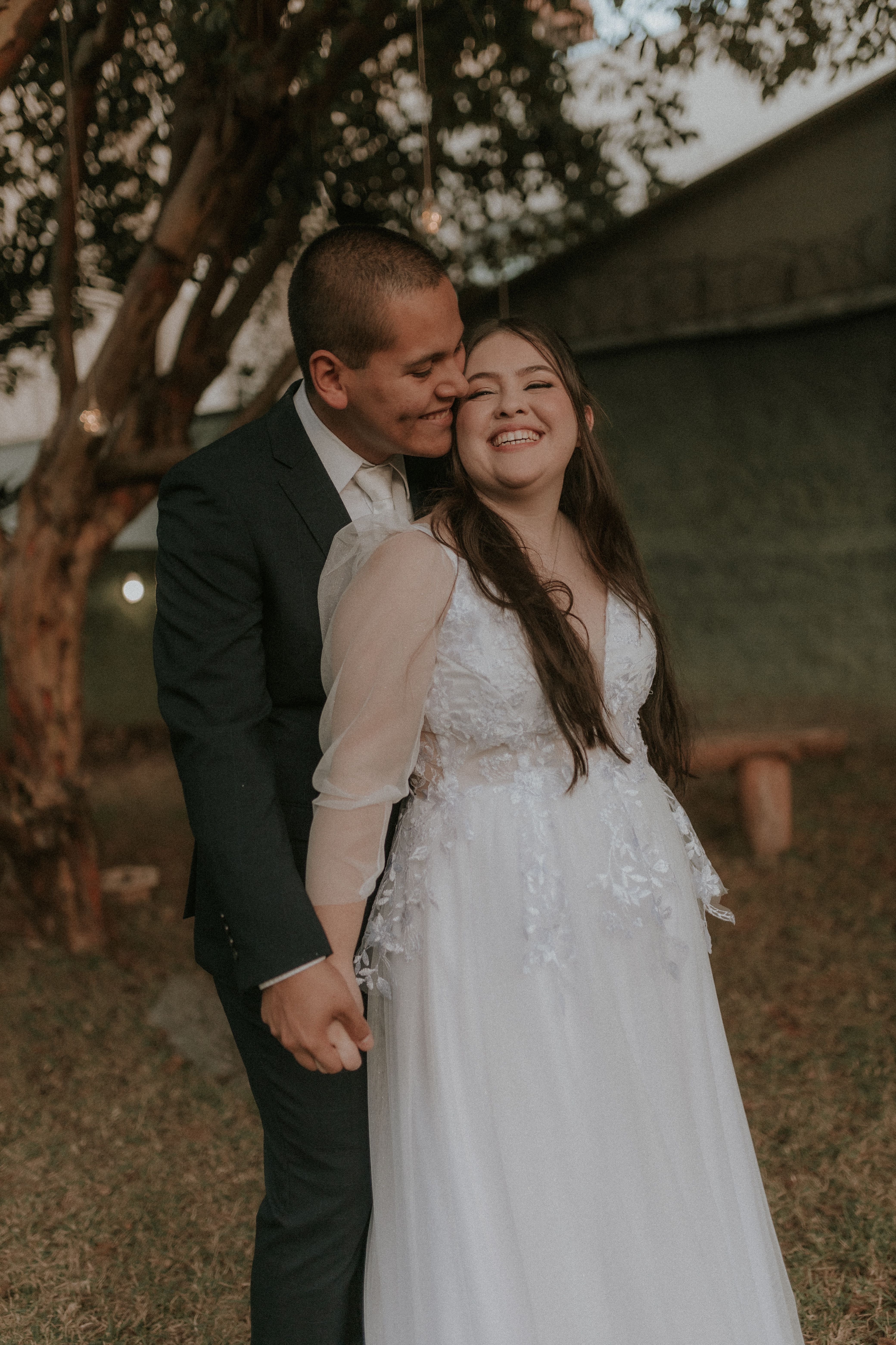 A bride and groom are hugging in front of a tree · Free Stock Photo
