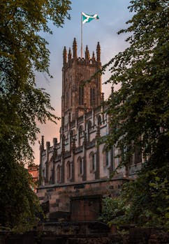 Majestic Gothic building in Edinburgh with a Scottish flag, framed by lush greenery. by David Rico