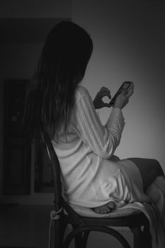 Artistic black and white portrait of a woman using a smartphone while sitting on a chair.