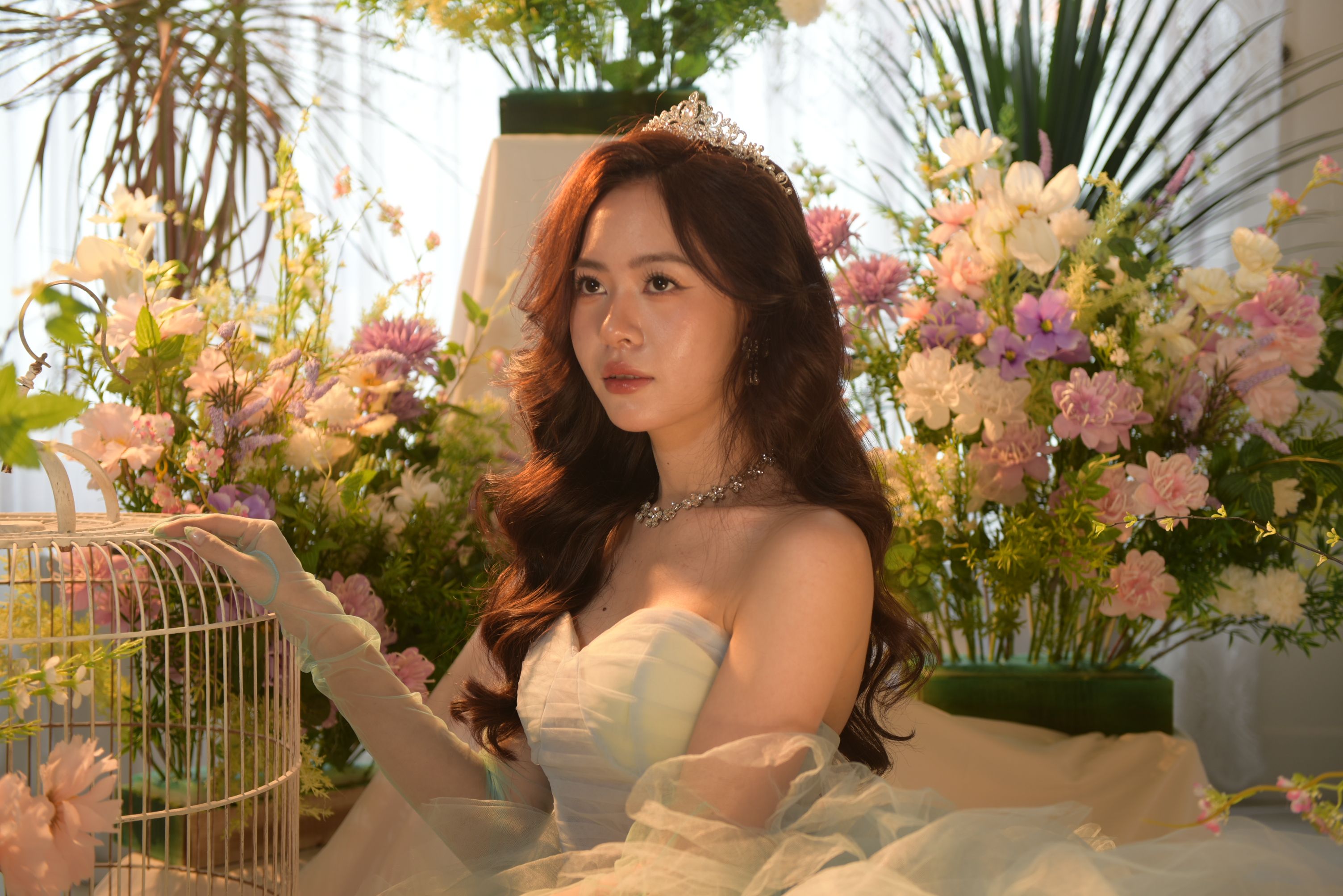 Beautiful Asian bride in a wedding dress, sitting among vibrant flowers in a sunlit room.