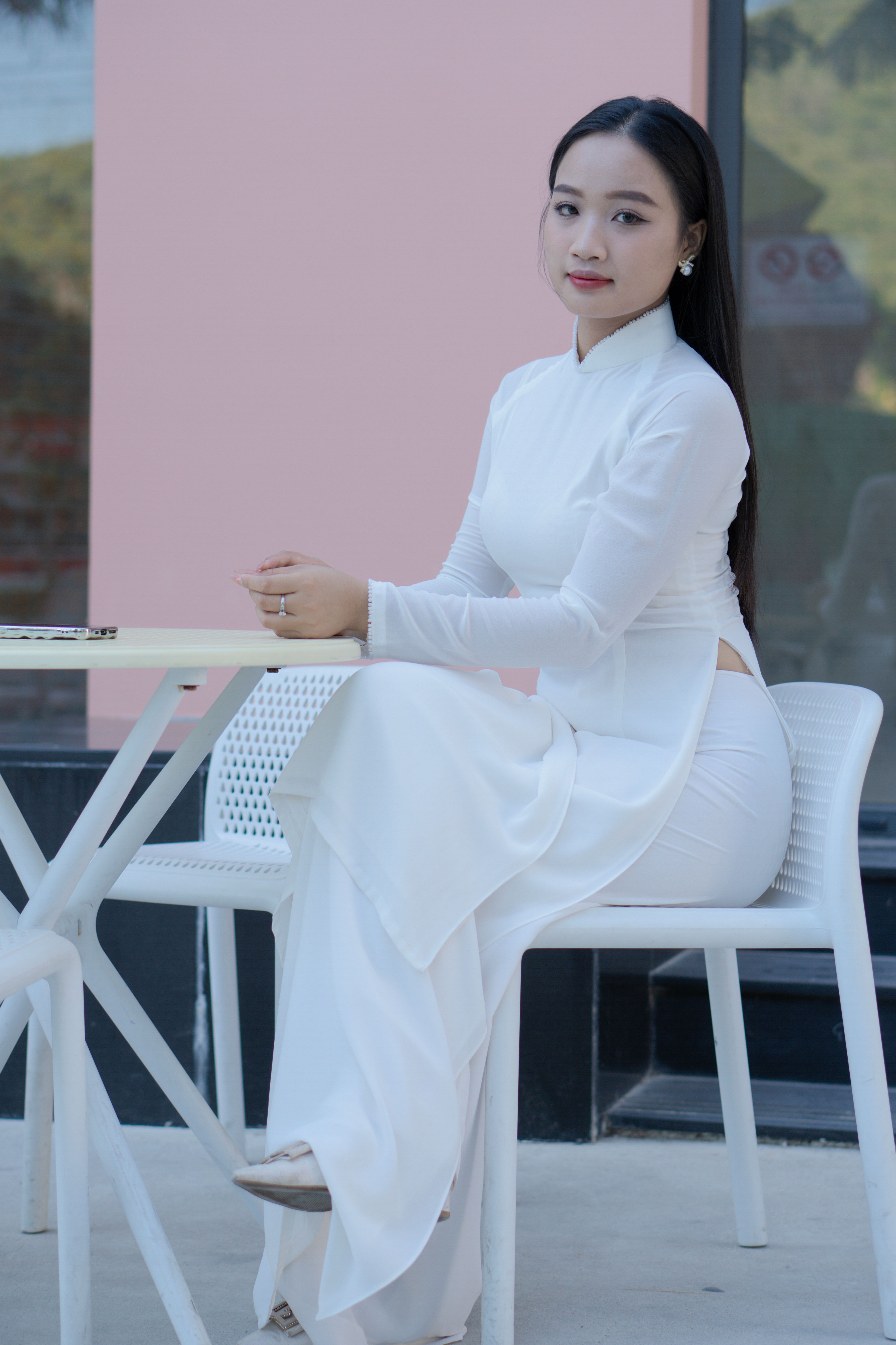 Young Vietnamese woman seated elegantly in traditional white Ao Dai, outdoors at a café table.