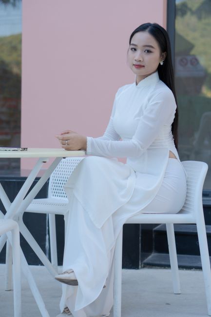 Young Vietnamese woman seated elegantly in traditional white Ao Dai, outdoors at a café table.