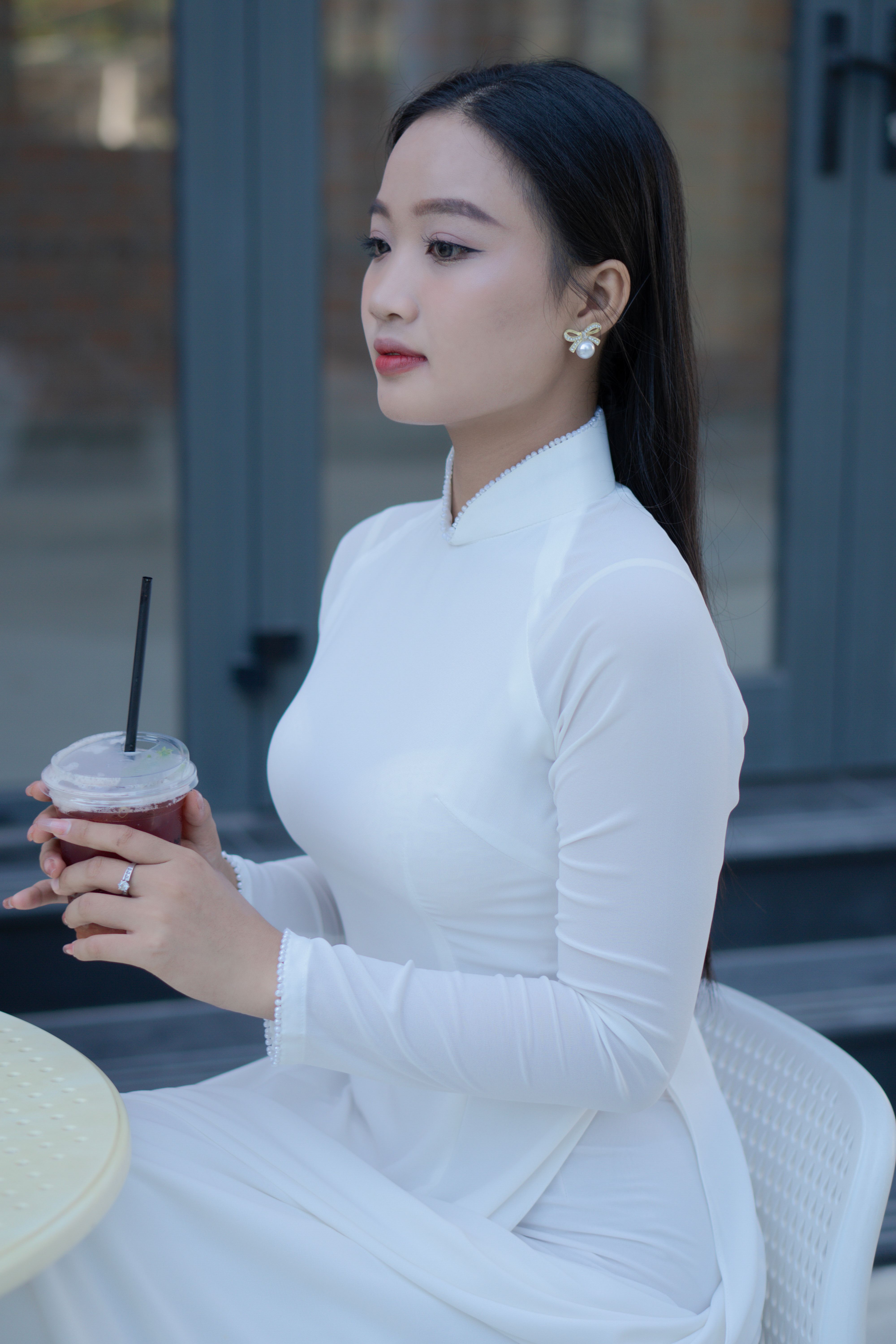 A serene young Vietnamese woman in a white Áo Dài sitting at an outdoor café.