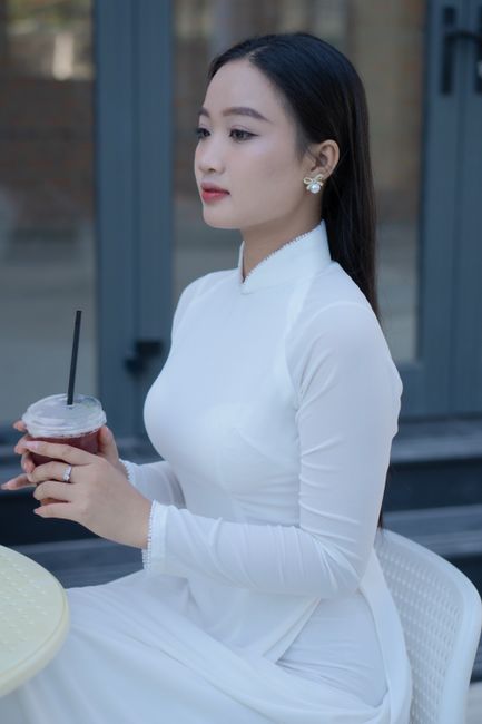 A serene young Vietnamese woman in a white Áo Dài sitting at an outdoor café.