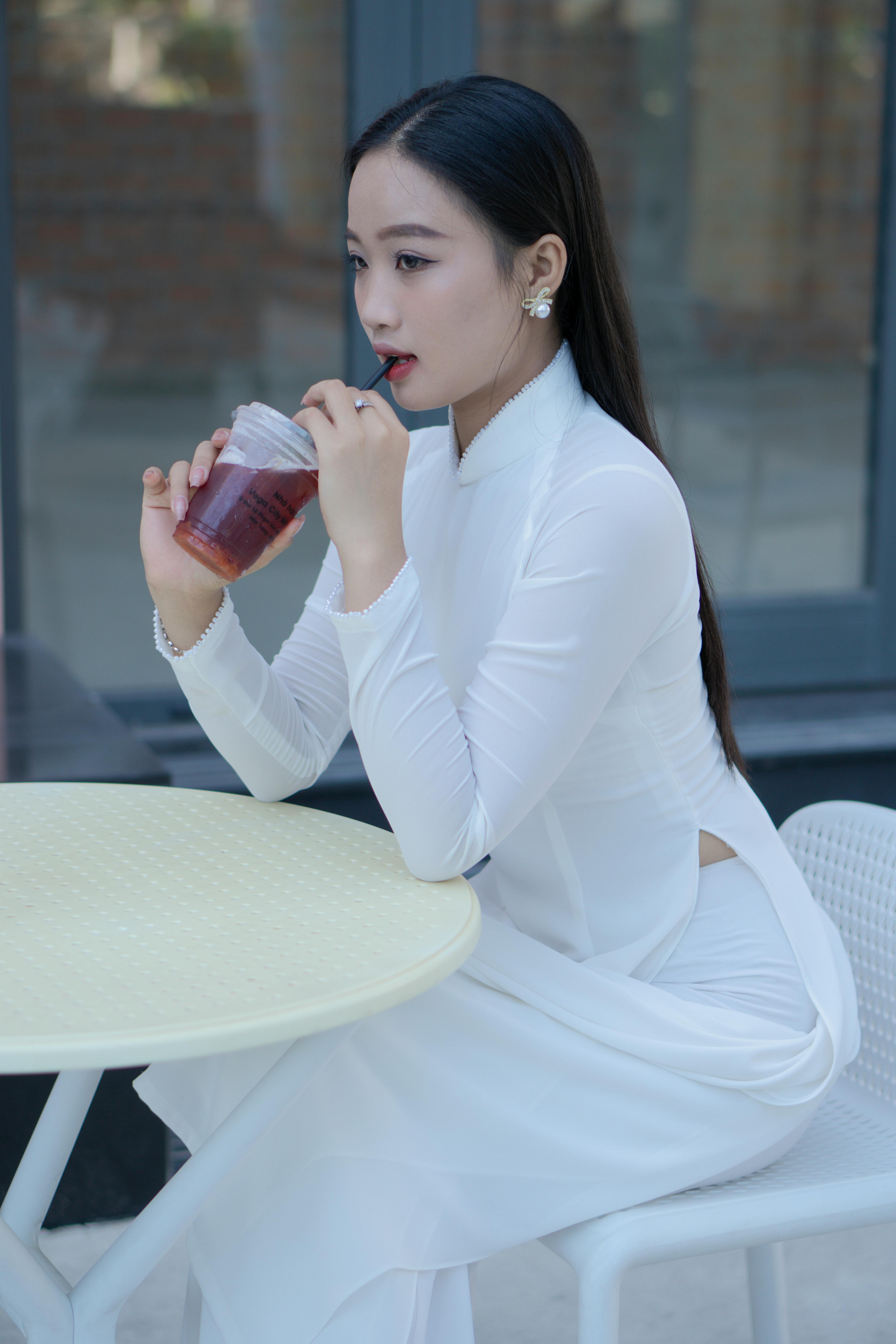 A young woman in a white Ao Dai enjoys a drink at a modern café.