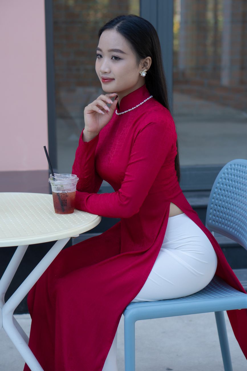 An elegant woman wearing a traditional ao dai sits outdoors, enjoying a drink and exuding confidence