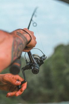 A detailed shot of a tattooed hand holding a fishing rod against a scenic backdrop in Italy.