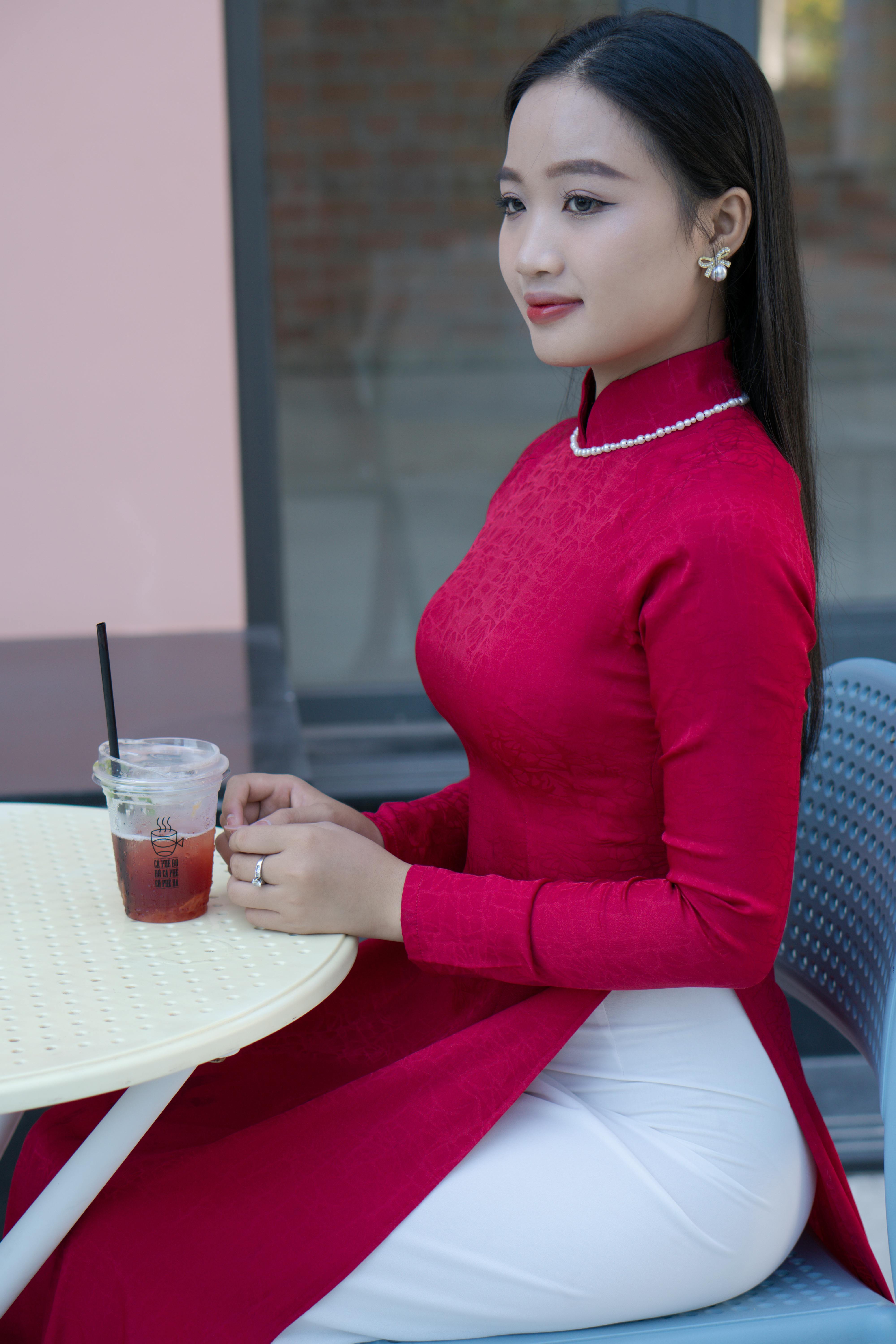 Young woman in red ao dai enjoying a drink at a cafe patio outdoors.