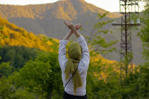 Free A woman wearing a hijab stands with raised arms amidst a picturesque mountain landscape. Stock Photo