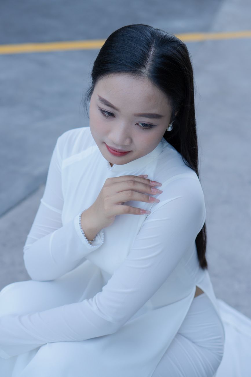 A beautiful Asian woman in a white ao dai dress sits gracefully outdoors.