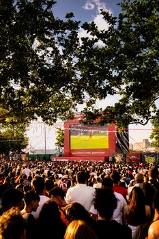 Large crowd gathered outdoors watching a football match on a big screen.