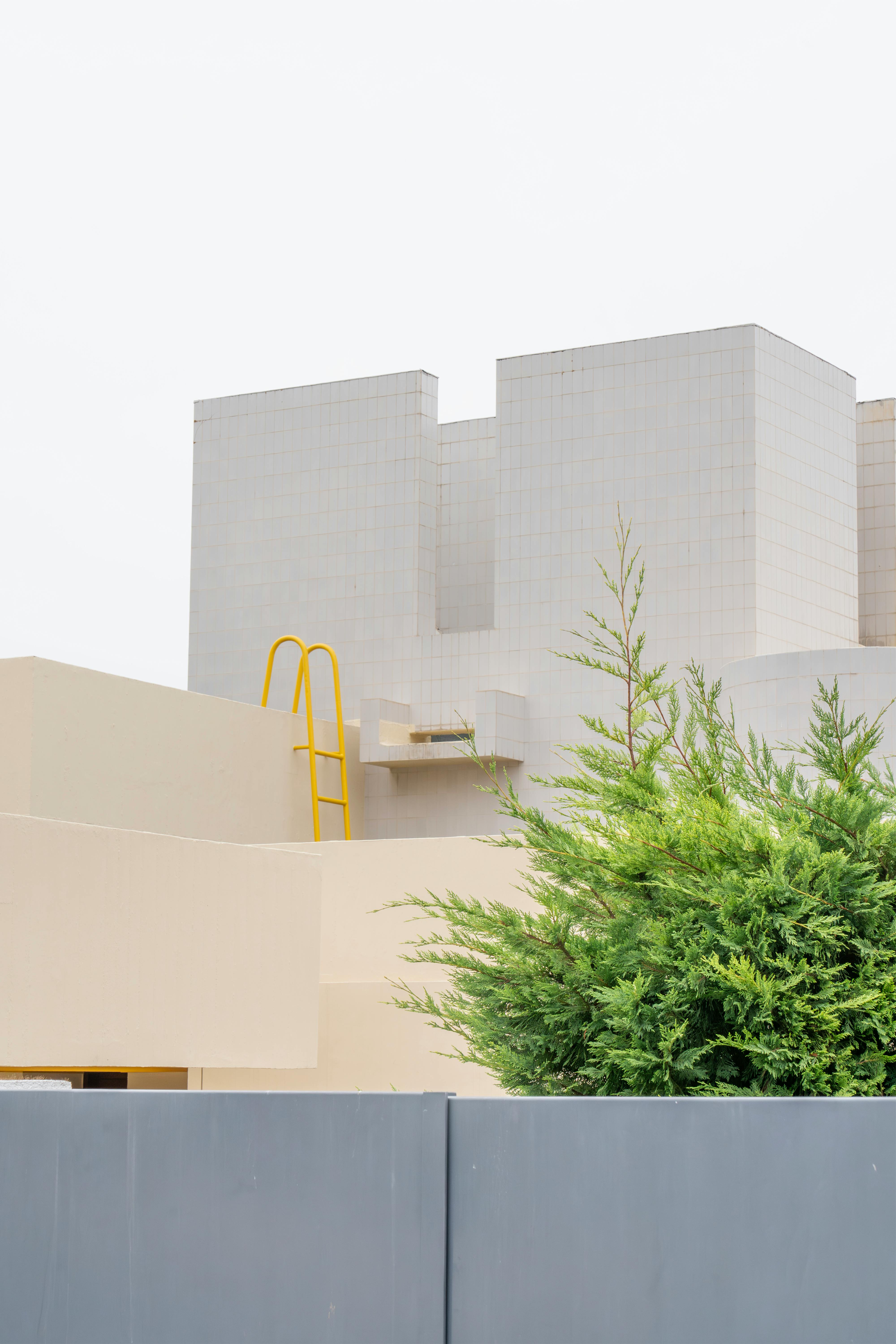 A minimalist modern building with a yellow ladder and cypress in the foreground, showcasing contemporary design.