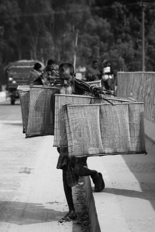 Free Black and white photo of an elderly man carrying large baskets in a city street setting. Stock Photo