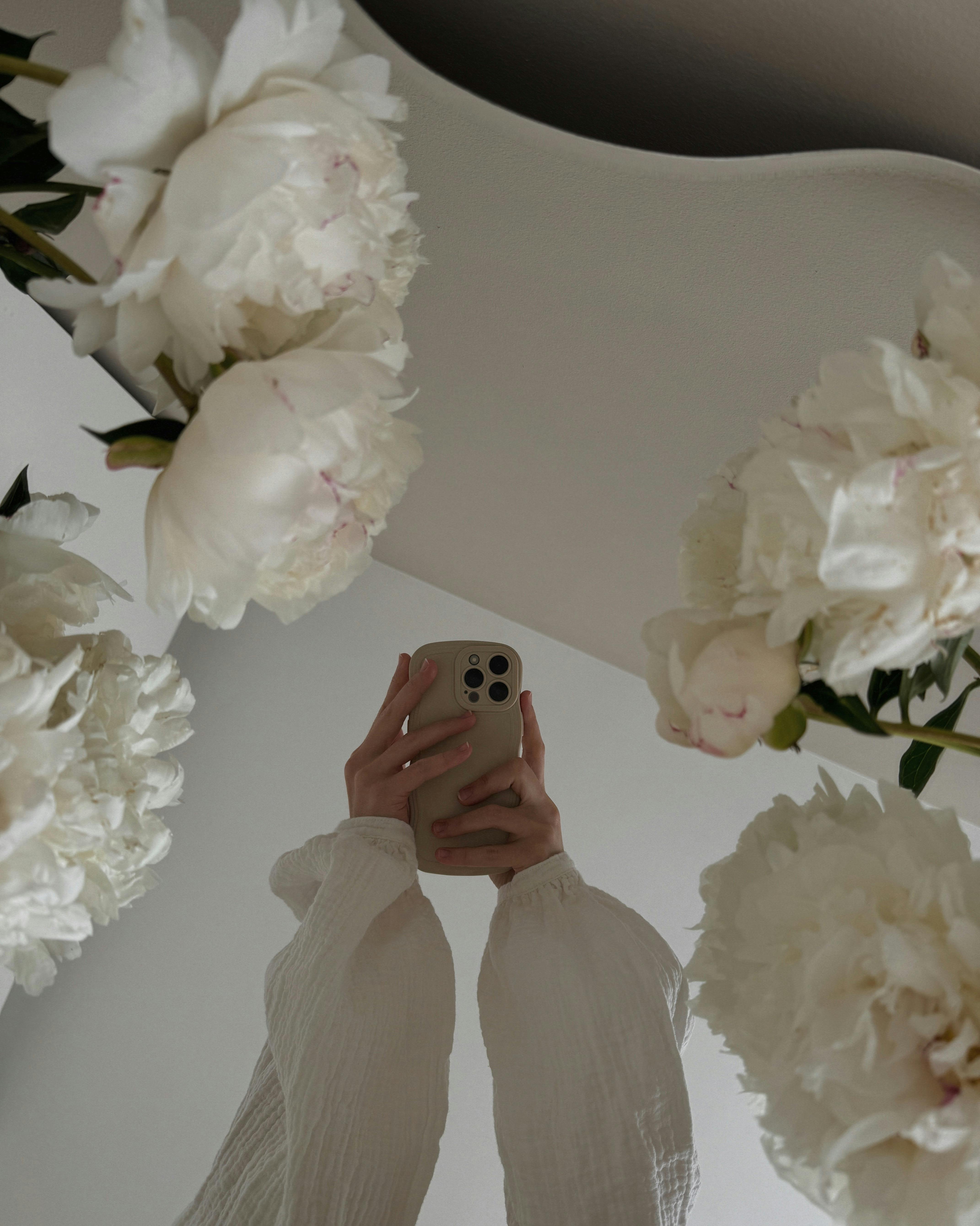 A serene reflection of a person holding a smartphone surrounded by white flowers in a mirror.
