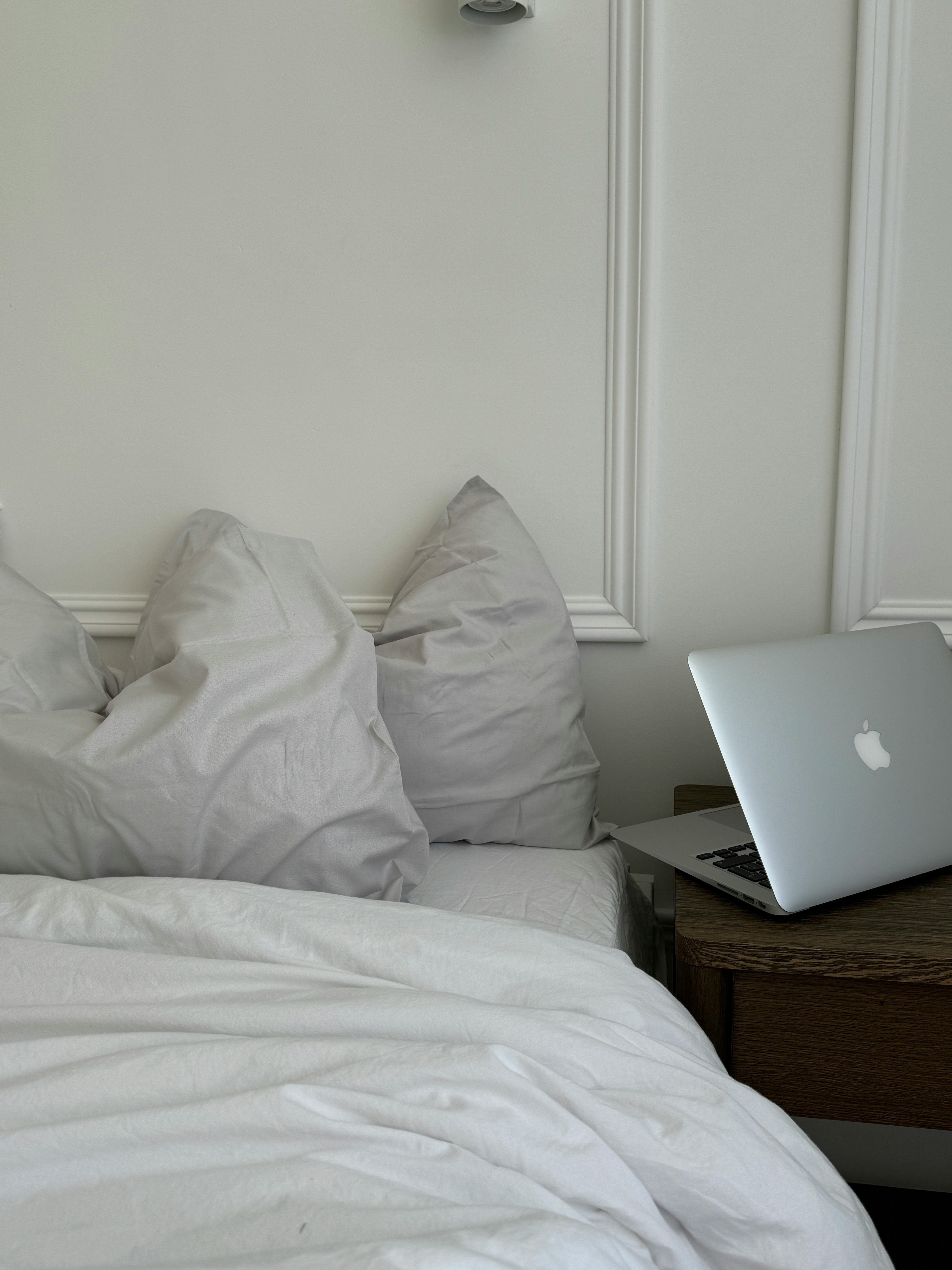 A cozy bedroom setting featuring pillows and a laptop on a bedside table, evoking a sense of relaxation and modern technology.
