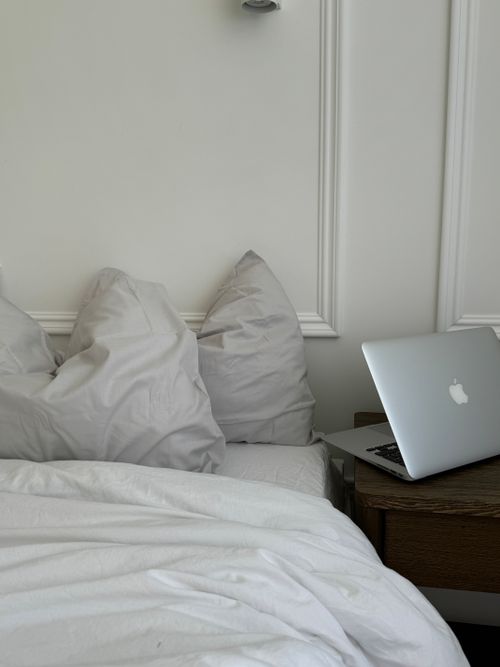 Free A cozy bedroom setting featuring pillows and a laptop on a bedside table, evoking a sense of relaxation and modern technology. Stock Photo