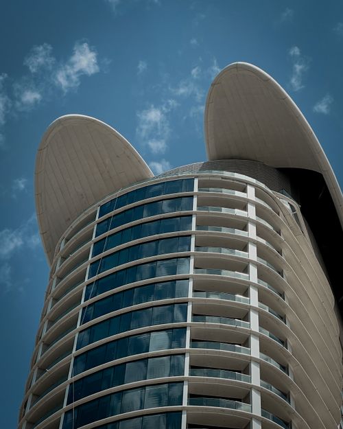 Free A sleek modern skyscraper with glass balconies and a unique curved roof against a blue sky. Stock Photo