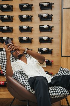 Man seated in modern chair using smartphone, surrounded by wine bottles on display.