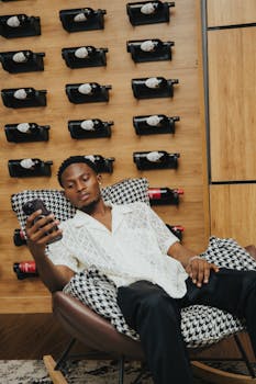 Man sitting in a chair, relaxing with a smartphone beside a wine wall in a modern setting.