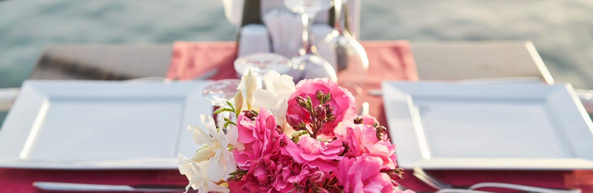 A beautifully set outdoor table with pink and white flower arrangement against a blurred sea backgro