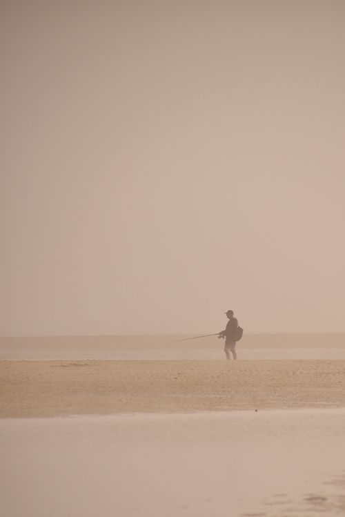 Free A lone fisherman stands on a foggy beach, casting his line into the calm sea. Stock Photo
